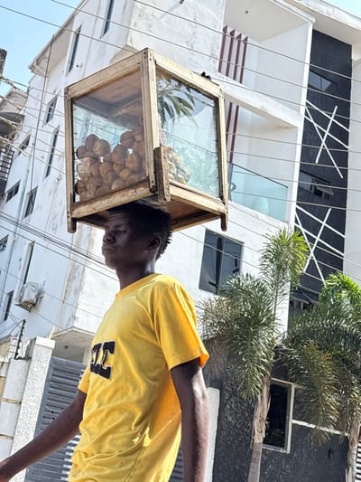 A male individual is observed in Igboefon, Nigeria, walking outdoors under bright daylight, balancing a rectangular display case on his head. The case is constructed from light-colored wood framing and glass panes, containing numerous round, light-colored food items, likely baked goods, on the left side, and smaller, irregular yellowish items, possibly fried snacks, on the right. The individual wears a bright yellow short-sleeved t-shirt with a dark, stylized logo on the chest. His head is slightly tilted backward, and his eyes are closed or squinted, indicating concentration on maintaining balance.
The background consists of two multi-story buildings. The structure to the left is predominantly white with multiple windows and an externally mounted air conditioning unit, partially obscured by a grey gate. The building to the right is a more modern design, featuring white and dark grey/black sections, accented with contrasting white vertical and diagonal line patterns. This building includes dark-tinted windows and a balcony with a glass railing. Several green palm trees are positioned in the mid-ground. Overhead, utility lines traverse the frame against a clear, light blue sky.