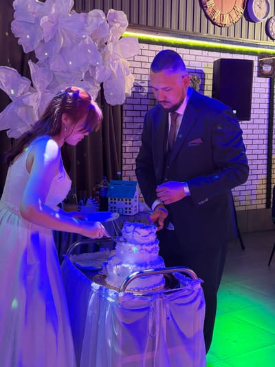 In Chernushka, Russia, at what appears to be a wedding reception, a woman in a white dress and a man in a dark suit are cutting a multi-tiered white cake. The woman is on the left, holding a knife and a plate, with her hair pulled back and adorned with a hair clip. The man is to her right, also holding a knife. The cake is decorated with white frosting and blue accents. They are standing behind a table draped with a white tablecloth. In the background, there are large white floral decorations, a brick wall, and some clocks on the wall. The lighting is a mix of blue and green, suggesting a festive atmosphere.