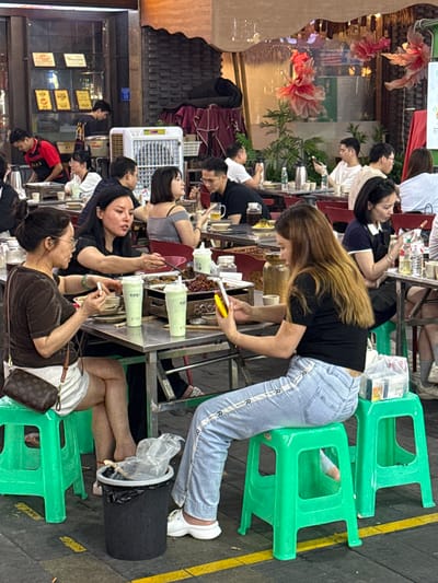 The image depicts a lively evening scene at an outdoor or semi-outdoor dining establishment, likely a street food market or restaurant alley in Liangjiang, China. The area is bustling with numerous people, primarily young adults, gathered around long, narrow tables.
The subjects include a multitude of diners, tables, and bright green plastic stools. In the foreground, a young woman with long, light brown hair, dressed in a black top and light blue jeans with a repeating 'R' pattern on the side, stands leaning over a table, holding yellow tongs. She appears to be serving or preparing food. Other individuals are seated, engaged in various activities such as conversing, eating, and looking at their mobile phones.
Notable details include the time of day, which is evening or night, indicated by the artificial lighting and overall ambiance. The setting suggests a casual, communal dining experience. The tables are filled with small dishes, food containers, and plastic cups holding a light green beverage. A white cooler or fan unit is placed between tables. In the lower right, a person's legs are visible next to a black cylindrical bin with a plastic bag liner, and a small brown handbag with a monogram pattern rests on a green stool. The background features decorative, illuminated red and pink floral structures and blurred storefronts with visible signs displaying Chinese characters. The overall atmosphere is vibrant and social.