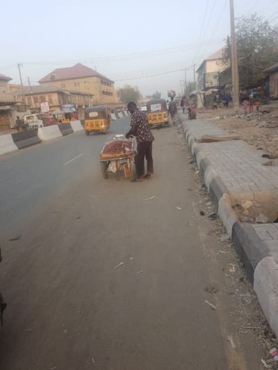 A man stands on a paved street next to a pushcart laden with peanuts. He wears a patterned shirt and dark trousers. A road stretches alongside, with yellow auto rickshaws and vehicles visible. Buildings stand on both sides of the street, and people walk in the background. Debris litters the edge of the road and the sidewalk. The air appears hazy.