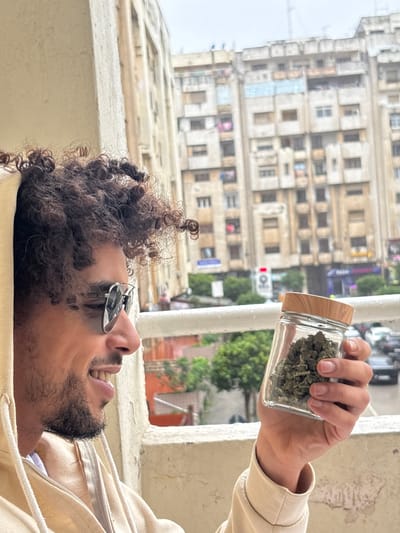 A person with curly hair and sunglasses smiles while holding a glass jar filled with cannabis buds. The jar is positioned near the person's head, against a backdrop of an old, multi-story building and some greenery. The individual wears a light-colored hooded sweatshirt. The photo seems to be taken from a low angle, possibly looking up at the subject.