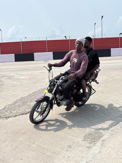 The scene in Igboefon, Nigeria, depicts two males on a black motorcycle on a light grey concrete road surface under bright daylight. The individual in the driver's position wears a light grey knit beanie, a plum-colored long-sleeved sweatshirt with a white 'B' logo, dark trousers, and black flip-flops. He grips the motorcycle's chrome handlebars. Seated directly behind him is a second male, attired in a dark short-sleeved t-shirt with vertical yellow embellishments, dark shorts, and blue and white patterned slides. The motorcycle features silver alloy wheels, a dark front fender with colorful decals, and a yellow rectangular reflective sticker on the front fork. No helmets are worn by either occupant. Distinct shadows are cast on the road. The background consists of a red barrier wall topped by a black and white striped barrier, above which a fence secured with coiled razor wire is visible. Two street light poles stand in the distance against a pale blue, hazy sky.