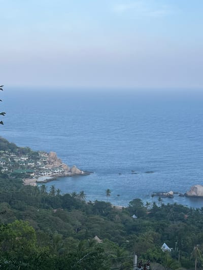 The image displays an elevated, panoramic view of a coastal area on Ko Tao, Thailand. The upper third of the frame shows a pale blue, hazy sky meeting a distant ocean horizon. The mid-section is dominated by the calm, blue-green sea, featuring subtle surface ripples and several scattered dark rock formations near the shore. The lower portion reveals a lush, mountainous coastline densely covered with tropical vegetation and numerous palm trees.
In the mid-left, a prominent rocky headland hosts a cluster of approximately fifteen to twenty white-walled buildings with light-colored or green roofs, resembling resort bungalows. These structures are built directly onto and around large, smooth grey boulders at the water's edge, framing a small sandy cove. Further along the coastline to the right, additional sandy beach segments are visible, backed by dense foliage and scattered structures, including at least one white conical-roofed building. No individuals or specific human activities are discernible within the frame. The lighting suggests either early morning or late afternoon.