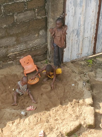 In Ukwu-Abwa, Nigeria, three children are engaged in play in a sandy area adjacent to a weathered brick wall and a corrugated metal door. A young girl stands in the background, dressed in a brown shirt and dark pants, leaning against the doorframe. In the foreground, two younger children are playing in the sand. One child, wearing a light-colored dress, is seated and appears to be handling sand. The other child is kneeling and interacting with an orange plastic toy, possibly a sand mold. An orange plastic chair, shaped like a bear's head, is placed near the children. Various objects, including what appear to be discarded items and a small figurine, are scattered in the sand. The environment suggests an outdoor play area, possibly a yard or communal space.