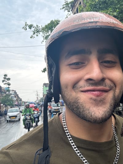 A medium close-up shot shows a man wearing a brown helmet with water droplets, a silver chain necklace, and a brown t-shirt. The man is looking at the camera with a slight smile. The background reveals a street scene in Kolkata, India, with cars, motorcycles, and buildings. The sky appears overcast.