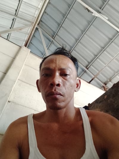 A man wearing a white tank top is pictured from the chest up. He is facing the camera, his expression is neutral. He has short, dark hair. The background shows a corrugated metal ceiling and a white wall. The scene is likely indoors or under a covered structure. There are no discernible landmarks to indicate the specific location within Bukit Ketok, Indonesia.
