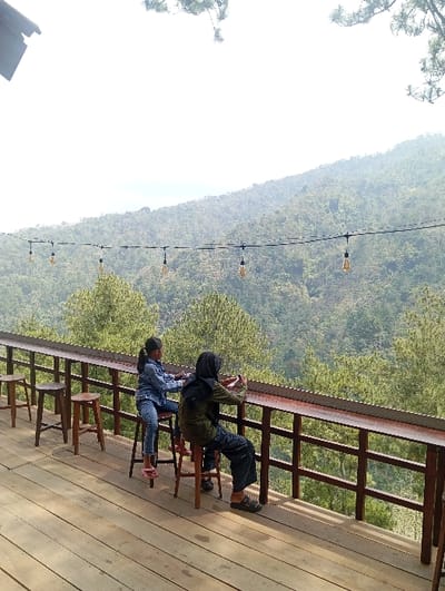 The image captures an outdoor viewing platform or cafe deck situated in the mountainous region of Kajar, Indonesia. Two individuals are seated on wooden bar stools at a long, elevated wooden counter that overlooks a vast, densely forested valley with rolling hills. The individual on the left, a younger person, wears denim attire and observes the distant landscape. The individual on the right, an adult wearing a dark headscarf and dark clothing, holds a white smartphone horizontally, pointed towards the scenic view, actively capturing content. The deck features light-toned wooden planks for flooring and a dark brown wooden railing system supporting the counter. Overhead, a string of several exposed-filament light bulbs is suspended across the frame. The background consists of an extensive, lush green forest under a bright, hazy sky.