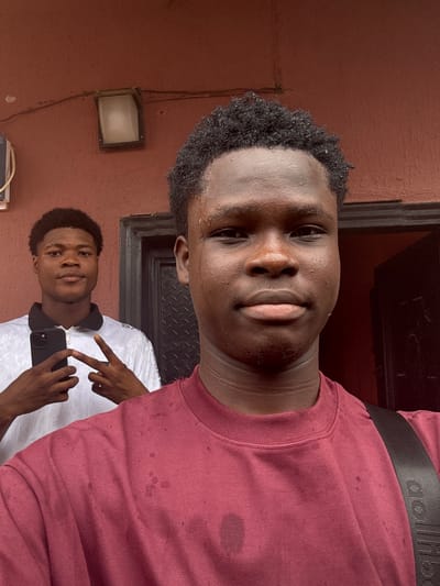 This image, captured in Ihiagwa, Nigeria, features two young Black males in an outdoor setting. The foreground subject, facing the viewer directly, has short, dark, curly hair with visible moisture droplets and wears a maroon crew-neck t-shirt displaying multiple darker spots consistent with sweat or water. A black strap, consistent with a bag or backpack, crosses his left shoulder (viewer's right). The second subject, a young Black male positioned behind and to the left, also faces the viewer. He is dressed in a white collared polo shirt with a black collar and zipper, holding a black smartphone in his left hand and forming a V-sign with his right. The background features a reddish-brown, textured wall. An outdoor light fixture with a rectangular frosted white cover and dark frame is mounted on the upper left, above which a black electrical wire runs horizontally. A dark, textured door frame and a section of a dark door are visible to the right. A portion of a light-colored utility box is visible on the far left.