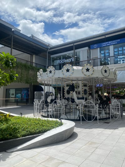 This image depicts an outdoor scene at a shopping complex in Baan Chaweng, Thailand. A white carousel with black and white animal figures, including horses and zebras, is the central focus. The carousel is adorned with lights and star decorations. In the background, there are modern buildings with storefronts displaying signs for "NUMAC STUDIOS FIXPHONE," "ISLANDS OF SIAM LUXURY REAL ESTATE LAND AND HOMES," and other businesses. A lush green plant bed with small yellow flowers is visible in the foreground. The ground is paved with light-colored square tiles. The sky is blue with scattered white clouds. There are no people visible in the immediate vicinity of the carousel.