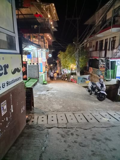 The image depicts a street scene at night in Bhagsu, India. The street is paved with concrete and lined with buildings on either side. The buildings appear to be multi-story structures with shops and cafes on the ground floor. There are several signs visible, including one for "Cafe" and another for "Tattoo Shop." A scooter is parked on the right side of the street. A few people are walking in the distance. Overhead, a complex network of electrical wires is visible. The overall atmosphere is that of a busy street in a developing urban area at night.