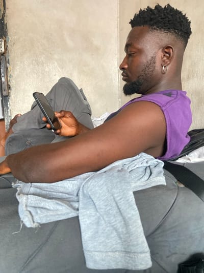 A dark-skinned man with an earring is sitting down looking at his phone. He has a fade haircut and a beard. He is wearing a purple shirt and grey pants. He is sitting on a grey couch with a grey shirt on it. The background is a white wall and a dark floor.