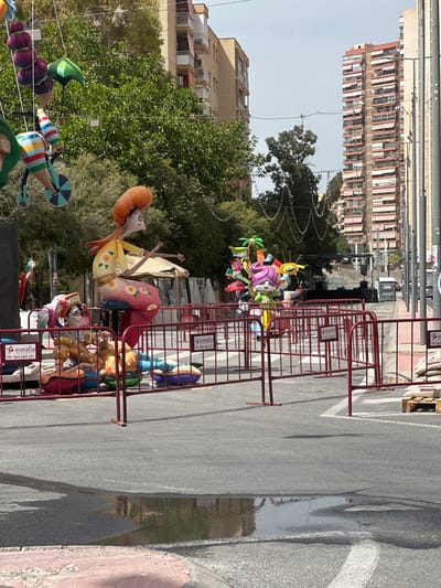 In Alicante, Spain, a street scene unfolds featuring vibrant, oversized figures reminiscent of Fallas sculptures, a prominent tradition in the Valencian Community. These artistic effigies, characterized by their exaggerated features and colorful designs, are displayed behind a red metal barricade. One figure, a woman with a distinctive orange, rounded hairstyle and a painted yellow and pink dress adorned with floral motifs, is seated. Beside her, another figure, wearing a checkered hat and a similar patterned outfit, is positioned.

The sculptures are set against a backdrop of lush green trees and the architecture of urban buildings. In the distance, tall residential buildings with numerous balconies and red awnings are visible. The street itself is paved, with a noticeable puddle of water reflecting the sky and surroundings. Various elements suggest preparation or a past event, including wooden pallets stacked with bags on the right side of the road and a portable toilet near a temporary stage setup in the background. Electrical wires crisscross above, strung between poles, and some decorative lights hang across the street. The overall impression is of an outdoor display in a public urban space, likely during a festive or commemorative period.