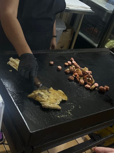 In a commercial kitchen, a person wearing a black t-shirt and black gloves is cooking on a large, flat griddle. The griddle is black and appears to be well-used, with some grease and food residue visible. On the griddle, there is a pile of sliced hot dogs and what looks like pieces of cooked chicken. The person is using a spatula to flip or move the chicken. To the left of the griddle, a stack of white paper towels is visible, along with a stainless steel shelf. In the background, through a glass-fronted appliance, a rack with heating elements can be seen. The setting suggests a food preparation area, possibly a restaurant or a food stall. The lighting is somewhat dim, and there are no clear indications of the time of day or weather. The overall atmosphere is one of active food preparation.