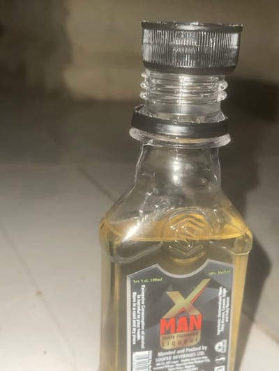 The image shows a close-up of a small glass bottle containing a yellow liquid. The bottle has a black plastic screw-on cap and a clear plastic neck with ribbed detailing. The label on the bottle is predominantly black with red and yellow lettering. The brand name appears to be "X MAN" with the tagline "Fruity Flavoured Liqueur." Other text on the label includes "Net Vol. 100ml," "30% Alc/Vol," "Ingredients: Demineralised Water, Ethyl Alcohol, Caramel, Sugar and Fruity flavour," and "Blended and Packed by SOOPER BEVERAGES LTD." There is also a warning about the excessive consumption of alcohol. The background is out of focus but appears to be a neutral-colored, possibly tiled or concrete floor. There are no people or discernible activities depicted in the image. The image does not provide any information about the location in the city of Howayi, Nigeria, or any other specific location.