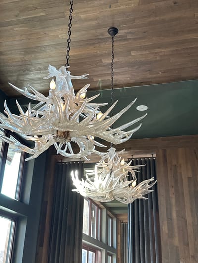 Two antler chandeliers hang from the ceiling, connected by chains. One chandelier is larger and closer to the viewer, while the other is smaller and further away. The background consists of wooden walls, a green accent wall, and windows with dark frames, allowing natural light to enter. The perspective is from below, looking upwards at the chandeliers.