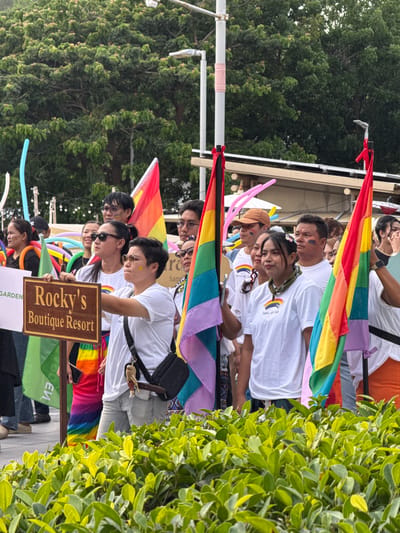 This media file captures a vibrant scene at a pride event, likely in Baan Chaweng, Thailand. A group of people, diverse in age and gender, are the central subjects, many adorned with rainbow-colored accessories, including flags, sashes, and face paint. They are seen standing together, holding rainbow flags, suggesting participation in a parade or celebration.
The setting appears to be outdoors, with lush green foliage visible on the right side of the frame, hinting at a tropical environment. In the background, hints of a structure, possibly a building or stall, can be observed. The time of day seems to be daytime, with bright lighting.
Notable details include the expressions on people's faces, which convey a sense of joy and pride. Some individuals are smiling, while others appear focused, perhaps on the event's proceedings. The presence of balloons in various colors adds to the festive atmosphere.
Visible text includes "Rocky's Boutique Resort" on a wooden sign, and the words "Samui HR Club" appear on some of the participants' white t-shirts, along with a rainbow logo. The overall mood is celebratory and inclusive, characteristic of a pride event. The activity depicted is the participation in and enjoyment of a pride parade or celebration.