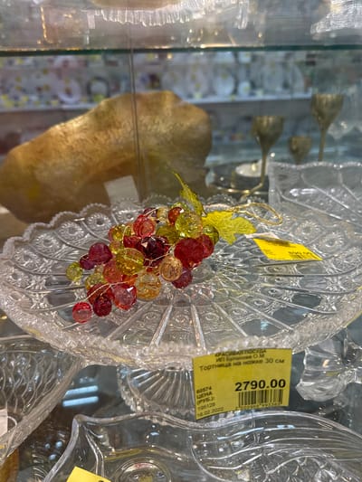 A clear glass cake stand adorned with a decorative cluster of red, orange, and yellow faux grapes with gold-colored leaves is displayed in a glass cabinet in a retail setting. A yellow price tag bearing the sum "2790.00" and text indicating "Тортница на ножке 30 см" (Cake stand on a stem 30 cm) and "ИП Буланова О.М." (IP Bulanov O.M.) is affixed to the cake stand. The date "16.02.2022" is also visible on the price tag. Based on the information on the price tag, it is assumed that this is a retail location in the Russian Federation. There is no indication that this is in the city of Klin, Russia.