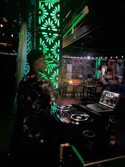 The image depicts a DJ performing in a dimly lit outdoor venue, possibly a bar or restaurant, in Howayi, Nigeria. The DJ, a Black male wearing a tie-dye t-shirt, is positioned behind a DJ setup featuring a laptop and mixing equipment, with several colorful buttons and lights illuminated. In the background, patrons are seated at tables, conversing and enjoying the ambiance. The venue is decorated with vibrant green floral patterns on vertical structures, and murals adorn the walls, adding to the lively atmosphere. A bottle of Budweiser is visible on the DJ's table, suggesting a social gathering. The overall scene conveys a sense of nightlife and entertainment.