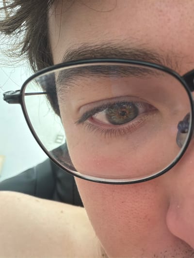 A close-up image focuses on a person's right eye, magnified through a lens of eyeglasses. The iris is visible, revealing green and brown tones. The eyebrow is dark and thick. The surrounding skin has fine lines and pores visible. The frame of the glasses is black. The background is indistinct, but appears to be a light-colored wall. It is difficult to identify the specific location within San Andrés Cholula, Mexico based solely on this image.