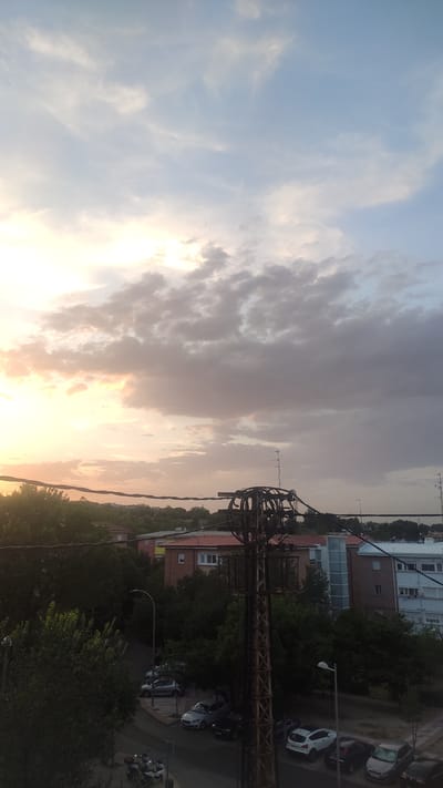 The image depicts a view of a residential area in Madrid, Spain, under a cloudy sky at dusk. In the foreground, a prominent rusty metal utility pole with numerous wires and electrical components stands on the right side, partially obscuring the scene. Beyond the pole, several residential buildings are visible, characterized by varying architectural styles and roof colors, including red and white. Trees line the streets and surround the buildings, creating a lush, green environment. A street is visible with parked cars, including a silver sedan, a white hatchback, and a dark-colored car. A streetlamp is also present. In the distance, the sky is filled with scattered clouds, with the setting sun casting a warm, orange and yellow glow on the horizon, transitioning to a lighter blue sky higher up. The overall impression is a serene, urban landscape during the evening hours.