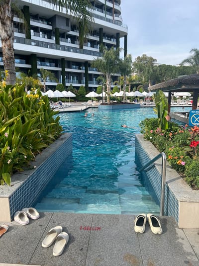 A resort pool area in Alanya, Turkey is depicted, featuring a large outdoor swimming pool with clear blue water. Steps lead into the shallow end of the pool, where four pairs of sandals are placed on the grey tiled edge. The pool is surrounded by lush green plants and flowering bushes. In the background, lounge chairs and white umbrellas are set up, with a modern multi-story hotel building towering behind them. Several people are visible in the pool, some swimming and others relaxing. A thatched-roof bar area is also present to the right, with a "No Diving" sign posted. The overall scene suggests a sunny day at a vacation resort.