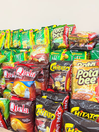 This is a vertical, eye-level, medium shot of a large display of potato chip bags, likely in a retail setting in Denpasar, Indonesia. The bags are stacked tightly together, creating a vibrant wall of packaging in various colors and designs.

The dominant brands visible are "Pota Bee," "Chitato," and "Qtela." "Pota Bee" bags feature yellow and red branding with images of chips and text like "Potato Chips" and "SAPIPANGGANG BBQ BEEF/BBQ." "Chitato" bags are predominantly dark brown or black with bright yellow lettering and images of chips. Some "Chitato" bags also show the text "BARBEQUE" and "RASA SAPI PANGGANG BEEF FLAVOR." "Qtela" bags are also dark brown with bold white and red lettering and feature images of chips and text such as "RASA ORIGINAL" and "RASA BARBEQUE BBQ Flavor." Additionally, some green bags with yellow text that appears to say "LITE" and "HANGOUt PACK" are visible, along with a smaller bag with text that seems to be "SINGKONG."

The image is well-lit, suggesting an indoor environment with artificial lighting. There are no people visible, only the merchandise. The sheer volume and arrangement of the snack bags indicate a commercial display designed to attract customers. The variety of flavors and brands hints at a general store, convenience store, or supermarket aisle in Indonesia. The overall impression is one of abundance and a wide selection of popular snack foods.