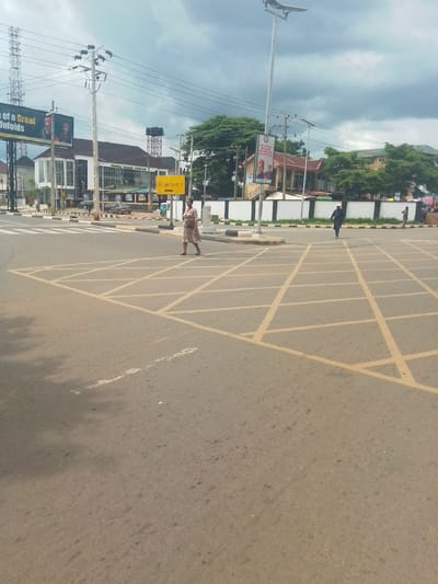 The image displays an urban street scene, likely an intersection or a wide road junction, captured on an overcast day in Awka, Nigeria. The foreground features a broad expanse of asphalt road marked with prominent yellow box junction lines.

Several individuals are visible. A woman wearing a patterned dress walks from left to right across the yellow road markings. Further back and towards the right, a man in dark attire and a white cap walks away from the viewer. Other pedestrians can be seen in the background near the buildings.

On the left side of the road, a modern white building with large glass windows and a dark roof stands out, bearing the inscription "ANAMBRA TRADE CENTRE." Behind it, a tall metal tower topped with a water tank is visible. On the far left, a tall lattice telecommunications tower rises into the cloudy sky.

The scene is rich with urban infrastructure. Numerous utility poles with a dense network of power lines crisscross the sky. Tall streetlights, some appearing to be solar-powered, punctuate the landscape. A large billboard on the left, partially reading "... OF A GREAT Unfolds," displays two male portraits, indicative of political advertising. Another smaller sign on the right features similar portraits and text including "...MAN THE PEOPLE TRUST... ANAMBRA NDIGBO... YOU CAN TRUST." A yellow directional sign points left, with the text "Enulu Agulu..." partially visible.

A few vehicles are parked or moving in the background. Green trees are interspersed among the buildings and along the road edges. The sky is heavily clouded, suggesting a dull, daytime atmosphere. White zebra crossing markings are also discernible further into the intersection.