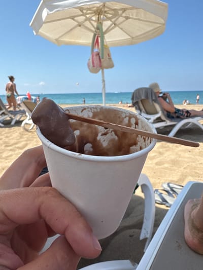 An outdoor medium shot captures a person's hand holding a white paper cup filled with melted chocolate ice cream on a sandy beach. A wooden stick extends from the cup, with a blob of chocolate ice cream on its tip. In the background, a white beach umbrella provides shade over beach chairs occupied by people. The sea stretches out beyond the beach, with a clear blue sky above. The overall scene suggests a relaxed day at the beach in Manavgat, Turkey.