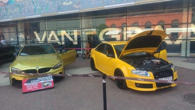 The image captures an outdoor event featuring two yellow sports cars parked on a brick-paved plaza in front of a building with large glass windows. The building's facade is decorated with a colorful mural depicting abstract floral motifs and trees. Visible through the glass is an interior space with brick architecture and large windows, suggesting a commercial or cultural establishment.
In the foreground, a bright yellow BMW M4 F82 is positioned to the left. It has its driver-side door open, and a red sign is placed in front of it. The sign displays text in English and Polish, indicating "CHARITY RIDES BMW M4 F82 Austin Yellow" and details about distance, speed, and a website address. The BMW has a black roof and dark-colored wheels.
To the right of the BMW is a bright yellow Audi sedan, possibly an RS4. Its hood is fully open, exposing the engine compartment. This Audi also features dark-colored wheels with red brake calipers visible. Both cars are roped off with a purple ribbon that has repeating logos and text. The ribbon is held up by black stanchions.
In the background, various bicycles are parked in front of the building, some of which appear to be part of the event display. Reflections of people and the surrounding environment are visible in the glass windows. The overall scene suggests a car exhibition or a charity event organized in a public urban space. The presence of "FUNDACJA PRZEJAZDY CHARYTATYWNE" (Charity Rides Foundation) on the ribbon and sign strongly suggests this is in Poland, and a quick search of the website on the sign confirms this is likely the Piotrkowska 217 Street exhibition in Łódź, Poland.