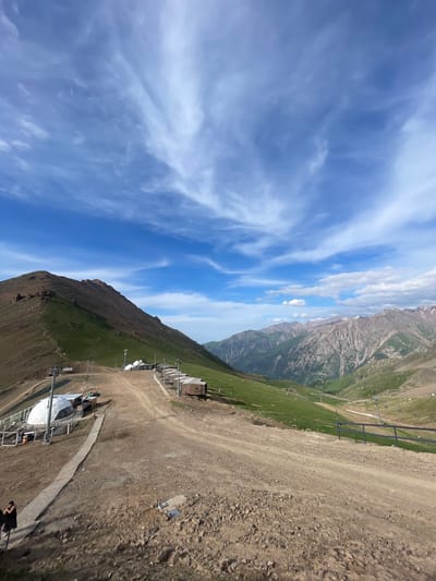 A wide-angle, high-angle shot captures a mountainous landscape under a partly cloudy blue sky. The scene is likely a ski resort or mountain recreation area. In the foreground, a dirt path curves uphill, leading past several small, shed-like structures and a large white dome-shaped building. A few people are visible on the path and near the structures. The path is lined with power poles and what appear to be ski lift infrastructure. To the left, a steep, grassy mountain slopes upwards, with rocky outcrops near its summit. In the background, a series of rugged, layered mountains stretch into the distance, some with patches of snow on their peaks. The overall environment is one of a high-altitude natural setting with developed recreational facilities. This scene is consistent with the mountainous terrain surrounding Almaty, Kazakhstan, particularly areas like Shymbulak or Medeo, which are popular for outdoor activities and offer similar views.