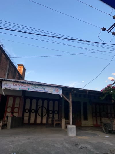 A daytime shot in Keudematangglumpangdua, Indonesia, shows a street scene under a bright blue sky with a few wispy clouds. Electrical wires crisscross in the upper left of the frame. On the right side is a building with a storefront or shop, partly obstructed by an advertising banner for "Berkah Laundry" hanging from the roof. The building has dark-painted, patterned doors. In front of the building is a concrete surface with a square concrete block. A portion of another building, likely made of brick, is visible on the left, with metal rebar protruding from the top.
