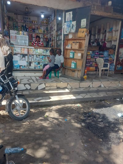 A street scene in Dar es-Salaam, Tanzania shows a storefront with various items for sale. A man and child are standing outside the storefront, interacting with someone inside. A motorcycle is parked in front of the store. A variety of goods are visible on the shelves inside the shop. The environment outside the store includes a dirt and paved sidewalk area. A plastic water bottle lays on the ground.
