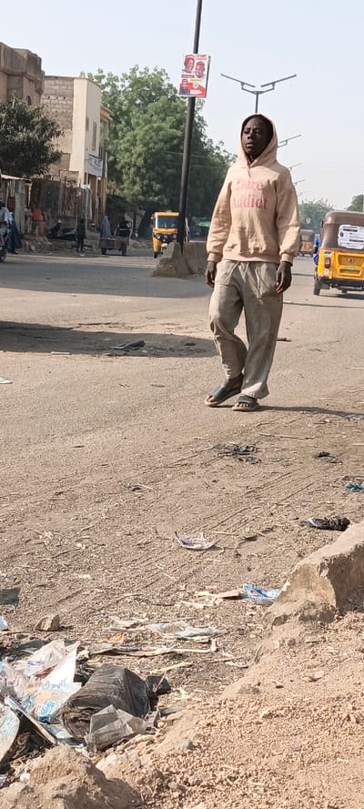 An outdoor street scene in Kaura Goje, Nigeria, is depicted under clear daylight. A dark-skinned individual, appearing male, stands in the center-right foreground, facing slightly left with a neutral expression. He wears a light peach-colored hooded sweatshirt with "Dare Addict" printed in reddish-brown text, loose light grey pants, and dark grey open-toed sandals.
The immediate foreground consists of rough, unpaved earth covered extensively with litter, including crumpled plastic bags (one black, one white/blue) and paper debris. Beyond this, a paved road extends into the midground, featuring concrete median barriers.
On the left side of the road, several indistinct figures are visible, along with a yellow three-wheeled auto-rickshaw. A person in a light-colored full-body garment pushes a two-wheeled cart along the road. On the far right, another yellow three-wheeled auto-rickshaw is present.
In the background, a line of buildings constructed from light-colored blocks and painted facades is visible on the left, interspersed with green trees. A dark street light pole stands to the left of the central individual, bearing a rectangular political poster featuring two smiling men in red caps and a red, green, and white national emblem. Additional streetlights with three-pronged fixtures are seen further down the road. The sky is clear and light blue.