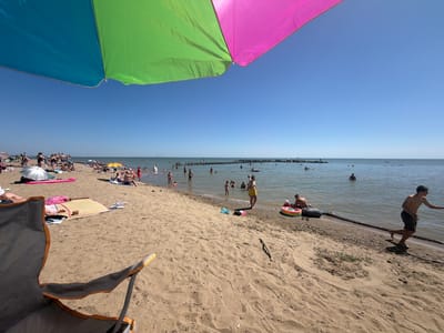 A high-angle, wide shot captures a sunny day at a sandy beach, with a large, multi-colored umbrella dominating the upper portion of the frame. The umbrella is divided into sections of blue, green, and pink, casting a shadow over the foreground. The beach is populated with numerous people engaged in various activities.
In the water, a crowd of individuals are swimming and wading, with some seen with inflatable rings. Several people are visible on the sand, some sitting or lying down on towels and mats, while others are standing. In the mid-ground, a group of people are gathered near the water's edge.
The environment is a typical beach scene, with sand, water, and a clear blue sky. The water appears calm, with a wooden structure visible in the distance. The overall impression is one of a leisurely summer day at a beach in Leninskoye, Russia.