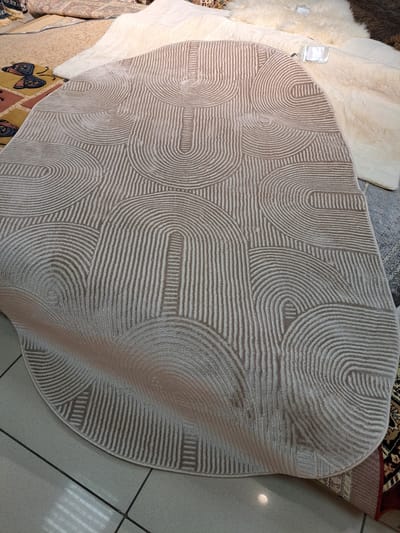 The image shows a beige rug with a geometric pattern of curved lines and rectangular shapes. The rug is partially unrolled and lies on a tiled floor. Several other rugs are visible in the background, including a fluffy white rug and a rug with a butterfly pattern. The scene appears to be a rug store or display area. The location is identified as Barnaul, Russia. There are no people visible in the image.