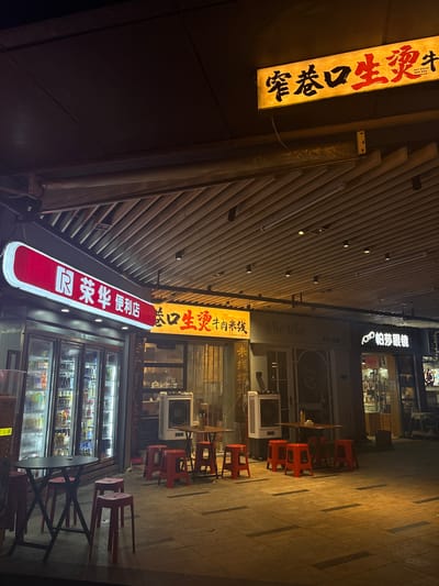 The image depicts a nighttime scene in the Haizhu District of China, featuring an outdoor seating area in front of several storefronts. On the left, a brightly lit convenience store, "R 荣华 便利店," is visible with glass-fronted refrigerators stocked with beverages. To its right, a restaurant with a prominent yellow illuminated sign displaying Chinese characters and the English text "HOT BOILED BEEF RICE NOODLES" is present. Adjacent to this is another establishment labeled "Via Via." Further to the right, a store with the sign "帕莎眼镜" is illuminated.
The ground in front of the stores is paved with tiles and is sparsely furnished with red plastic stools and a few small tables. Two portable air coolers are positioned in front of the restaurant area. The overall environment suggests a street or promenade with commercial activity. No people are visible in the image. The lighting is artificial, casting shadows and highlighting the signs and illuminated interiors.