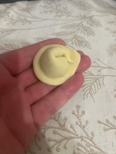 A close-up shot shows a person's hand holding a single, pale yellow, uncooked dumpling. The dumpling is round with a crimped edge and a twisted top, resembling a tortellini or a similar filled pasta. The hand is pink and appears to be resting on a textured, light-colored surface with a subtle pattern of faint, beige branches or leaves. The background is softly blurred, suggesting a shallow depth of field, and the lighting is even. There is no information in the image to suggest a specific location in Anapa, Russia.