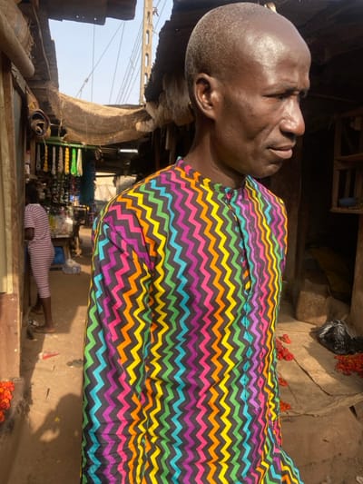 An elder African man with a shaved head is photographed outdoors, wearing a shirt with a chevron pattern of black, orange, yellow, green, blue, and purple. He's positioned at an angle, looking to the left. The background shows a market scene with various goods displayed, including shoes. Another person in a striped outfit lies on the ground. Additionally, there are red fruits or vegetables scattered around on the ground. The scene suggests a bustling, everyday life in an African marketplace.