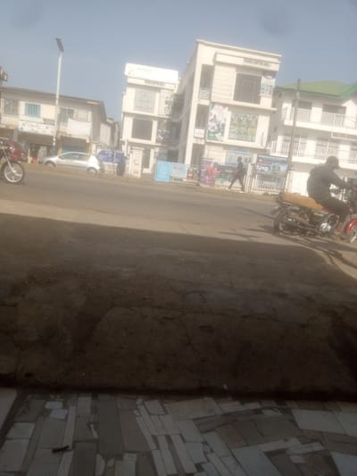 The image depicts a street scene in Dadi Riba, Nigeria, under a clear, bright sky. In the foreground, a dirt path or road occupies the lower portion of the frame, with some indication of paving stones or a tiled surface at the very bottom. Across the street, a modern, multi-story building with a white facade is visible, labeled "IBARRAKA SHOPPING MALL 1". Adjacent to it, another building, possibly an older structure, features a light-colored facade and some blue window frames.
Several individuals and vehicles are present. A person is riding a red motorcycle in the middle ground, moving away from the viewer. Another person, wearing dark clothing and a baseball cap, is also on a motorcycle, partially visible. A silver car is parked on the side of the road. Two pedestrians are walking on the sidewalk in front of the shopping mall. Various signs and advertisements are displayed on the buildings and on the street, including posters for "Sambo" and other commercial promotions. The overall environment appears to be an urban or semi-urban setting with commercial activity.