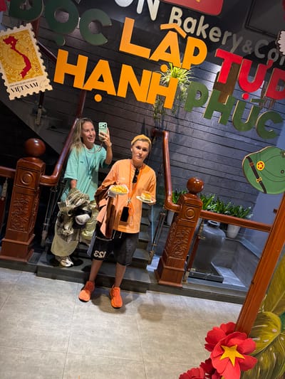 Two individuals are positioned on a wooden staircase with a grey-tiled floor. The person on the left, wearing a teal t-shirt, is holding a smartphone and taking a selfie. They are carrying a plaid shirt and a cap. The person on the right, in an orange t-shirt, holds a plate with two pieces of cake. They are wearing black shorts and orange sneakers. A large wooden railing with carved details runs along the staircase. Above them, large, colorful letters spell out "C LAP TU HANH PHUC" on a dark, textured wall. A stamp-like graphic of Vietnam is visible on the left side of the wall. Further up the stairs, potted plants and a large dark vase are present. The scene appears to be a well-decorated indoor space, potentially a cafe or restaurant, in South Quy Nhon Ward, Vietnam, given the visual cues.