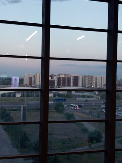 The image captures a wide shot of a cityscape as viewed through a large window, divided by dark vertical and horizontal mullions. The scene depicts a modern urban environment with a twilight sky, suggesting either early morning or late evening. Several large, multi-story buildings, likely residential or office complexes, dominate the middle ground. Some of these buildings exhibit a uniform architectural style with rows of windows. To the left, a distinct, tall building with a luminous, purple facade stands out.

In the foreground, a road with several vehicles is visible, including a blue bus and at least one white bus, along with cars. The presence of these vehicles suggests ongoing traffic and activity. The ground surrounding the road appears to be a mix of paved areas and undeveloped, grassy land with scattered trees. The overall setting is an urban landscape in Astana, Kazakhstan, characterized by contemporary architecture and infrastructure.

The lighting and soft colors of the sky indicate a transitional period of the day, likely dusk or dawn. There are no discernible people in the image, and the focus is on the built environment and the transportation infrastructure. The mood is calm and observational, offering a detached view of the city. No visible text is present in the image.