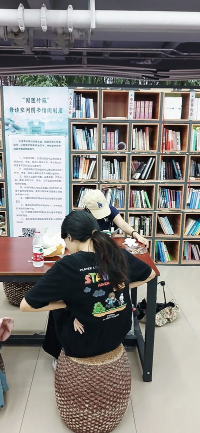 The photo shows a scene inside a library or reading room, possibly located in the High-tech Zone of Chongqing, China, given the presence of Chinese text on signs and books.

Two young women are present. One woman is standing at a table, reaching towards a shelf of books. She is wearing a beige cap and a dark top. The second woman, wearing a black t-shirt with printed designs and the words "PLAYER 1," is seated on a brown woven stool, possibly leaning over the table. She has long, dark hair.

Behind the women is a wall of bookshelves filled with books. A poster or sign in Chinese is placed in front of the bookshelves, likely containing library rules or information.

On the table, there are some items, including what appears to be a bottle of water and a container of food. A shopping bag or cloth bag is sitting on a shelf next to the book shelves.

The room is brightly lit and has a clean, modern appearance.