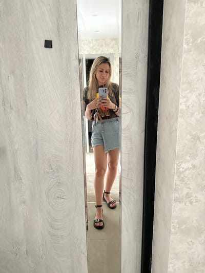 A woman is taking a selfie in a full-length mirror. She is wearing a dark t-shirt, light denim shorts, and black sandals. She is holding a purple smartphone in her right hand and a set of keys with a charm in her left hand. The mirror is framed by a black border and is situated in a room with light-colored, wood-patterned walls. The floor is tiled. Based on the available information, the location is the city of Novoaltaysk, Russia.