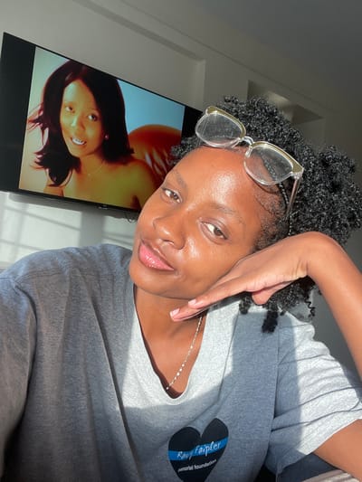 This image captures a close-up, indoor portrait of an adult female with dark skin in Thika, Kenya. She wears a grey short-sleeved t-shirt with a blue heart logo containing text "Randy Faipler memorial foundation" on the left chest. Clear-framed eyeglasses rest on her head, and a thin silver-colored necklace is visible around her neck. Her left hand is positioned with fingers resting against her jawline as she looks directly towards the viewer with a neutral, slightly smiling expression. In the background, a flat-screen television is mounted on a light-colored wall, displaying an image of another dark-skinned female with dark hair, smiling and looking upward. Strong, directional natural light illuminates the scene from the left.