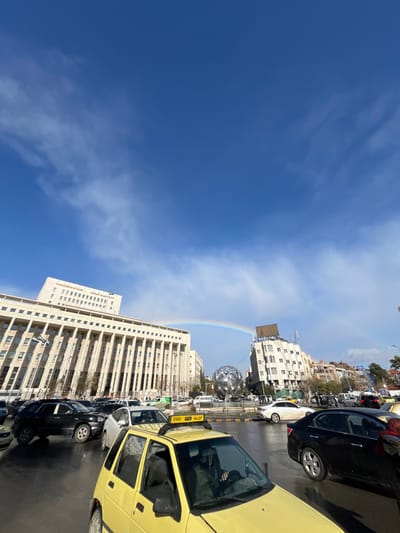 The image depicts an urban street scene in Sarouja Municipality, Syria, under a clear blue sky with a visible rainbow arc. In the immediate foreground, a bright yellow taxi, possibly a Daewoo Matiz or similar compact model, is oriented towards the right, with its driver partially visible behind the wheel, wearing a dark green garment. The taxi roof features a yellow sign displaying "TAXI" in English and "اجرة" in Arabic. The street surface appears wet, suggesting recent rain.

In the midground, a traffic lane is visible with multiple vehicles, including a black SUV, several sedans (white and black), and other cars, suggesting a flow of traffic or a stopped queue. Central to the scene, in what appears to be a roundabout, stands a large, metallic, multifaceted spherical sculpture.

The background features prominent urban architecture. To the left, a large, multi-story building with a classical facade characterized by numerous columns is identified by signage as the "Central Bank of Syria" in both English and Arabic. To the right, additional multi-story buildings of varying designs line the street. The rainbow spans across the sky, arcing from above the Central Bank building towards the right side of the frame, passing above the metallic sphere and midground buildings. The overall environment indicates daytime with bright illumination.