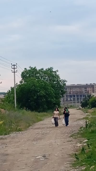 Two women walk down a dirt road in Baksan, Russia. The woman on the left wears a light-colored shirt and dark pants, carrying two plastic bags in her right hand. The woman on the right wears a dark shirt and dark pants, carrying two plastic bags in her left hand. A large green tree stands to the left of the road. In the background, an unfinished building with multiple stories is visible, along with power lines and a utility pole. The sky is overcast.