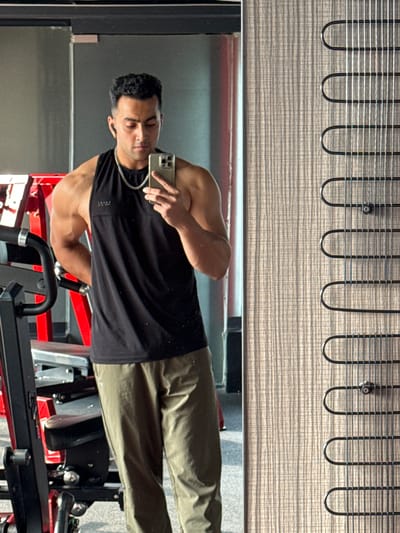 The image appears to be a mirror selfie taken in a gym setting. A man with short, dark curly hair, wearing a black tank top and olive green pants, is holding a phone and looking at his reflection. He is also wearing a chain. The gym equipment is partially visible behind him. The background features what seems to be a window shade at the top of the reflection. It is not possible to determine the location as Kolkata, India, without additional information.