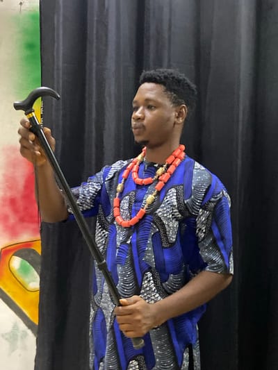 A young adult male, appearing to be of African descent, is positioned in an indoor setting in Mararaba, Nigeria. He has short, dark, textured hair and a subtle mustache and beard, with his gaze directed slightly upwards and to the left. He is wearing a short-sleeved tunic patterned with blue, grey, black, and white motifs, consistent with African print fabric. Around his neck, he wears a multi-layered necklace of large, cylindrical red coral beads interspersed with gold-colored, ornate spherical and cylindrical beads. He holds a black walking stick with a curved handle; his left hand grips the upper portion of the shaft below a gold-colored collar, while his right hand holds the lower shaft. The background on the left reveals a section of a painted wall or mural featuring segments of green, red, and yellow, with a distinct stylized eye shape visible. The background to his right and directly behind him consists of a dark, vertically folded curtain, likely black or dark grey. The scene is illuminated with even, diffused lighting.