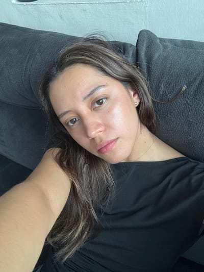 A medium shot captures a woman of Latin descent lying on a dark gray textured couch. The woman is looking directly at the camera with a neutral expression. Her long, dark brown hair is parted in the middle and falls over her shoulders. She is wearing a black, off-the-shoulder top and a delicate gold necklace. A small gold earring shaped like a crescent moon is visible in her left ear. The background consists of a textured white wall. The lighting suggests an indoor setting, likely during the day, with soft light illuminating her face. The image appears to be a selfie. There is no indication of the specific location within Puebla, Mexico.