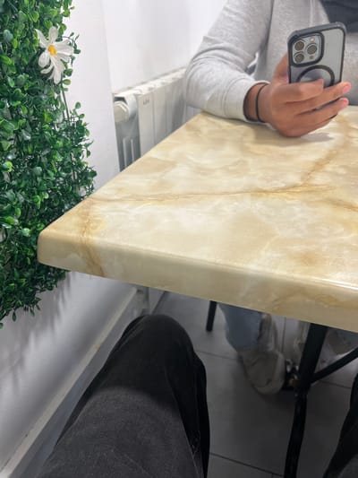 The image shows a person holding a smartphone to photograph what appears to be a countertop or surface with a faux marble pattern. The countertop is positioned above a decorative green plant with a white flower. A radiator is visible behind the plant.
In the background, there is a person wearing dark pants and a chair with black legs. The location seems to be indoors, with white walls and tiled floors. The city is not identifiable based on the image alone.
